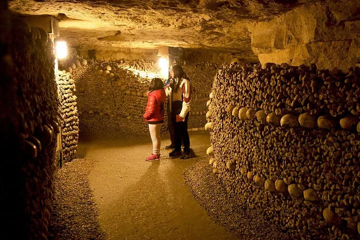 Two people standing in a dimly lit tunnel viewing walls made of bones.